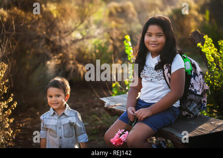 A boy and his older sister are sitting down, taking a break during a hike or field trip while the sun is setting. Stock Photo