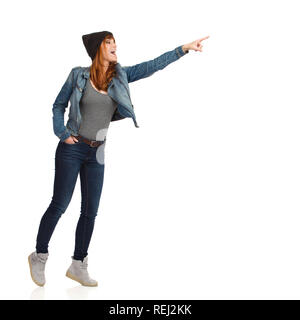 Young woman in jeans jacket and cap is standing tip toe, pointing, looking away and laughing, Full length studio shot isolated on white. Stock Photo