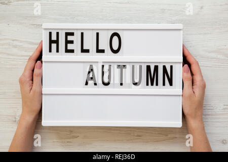 Female hands hold lightbox with text 'Hello Autumn' over white wooden background, top view. Overhead, flat lay, from above. Stock Photo