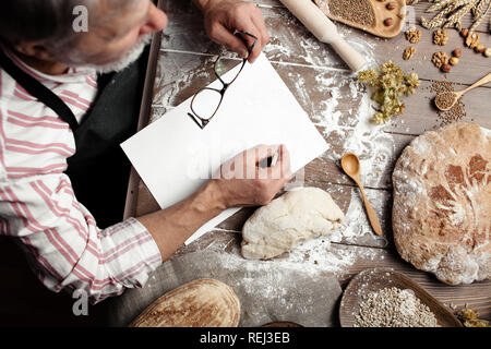 Old Baker writing down old-time recipe in bakery notebook surrounded by bread Stock Photo