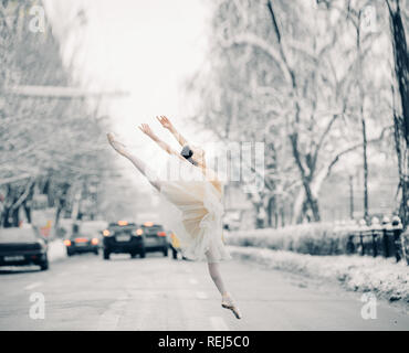 Beautiful ballerina in transparent skirt is dancing and jumping on street of snowy city among cars. Stock Photo