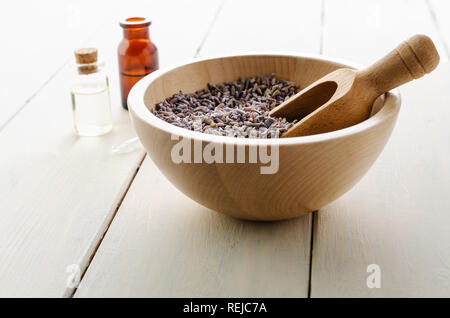 Herbal Lavender flower buds fill a wooden bowl with scoop in front of glass vials containing oils with pipette on white painted wood plank table. Stock Photo