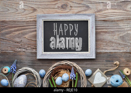 Easter black board with Easter eggs, flowers and spring decorations on rustic wood, text "Happy Easter" on the board. Stock Photo