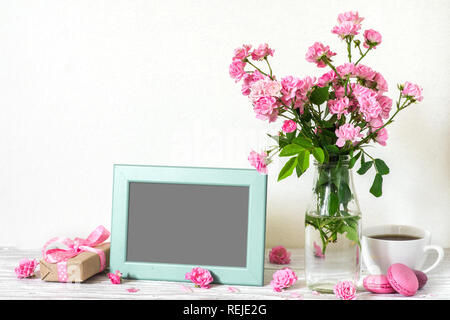 blank photo frame with beautiful bouquet of pink roses flowers, coffee cup, macaroons and gift box. holiday or wedding background. mock up. still life Stock Photo