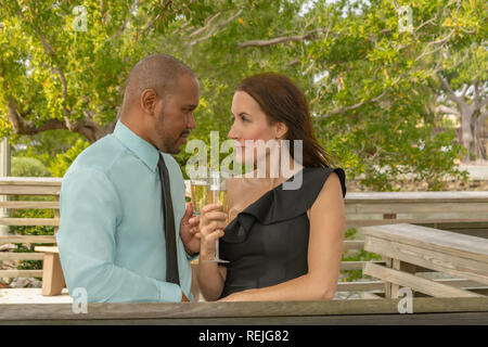 A multiracial couple gazes into each other's eyes while toasting with champagne. Enjoying the moment celebrating outdoors on a deck. Stock Photo