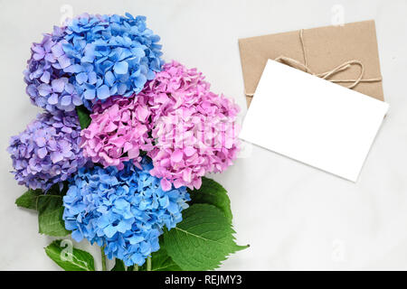 blank white greeting card with hydrangea flowers bouquet over white marble table with copy space. top view. mock up. wedding invitation Stock Photo