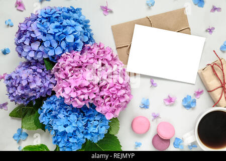 blank greeting card with pink and blue hydrangea flowers, coffee cup, gift box and macaroons over white marble table with copy space. top view. mock u Stock Photo