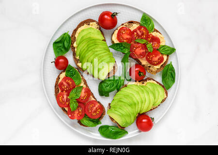 healthy breakfast toasts with avocado, tomatoes, hummus and basil over white marble table. vegan food. top view Stock Photo