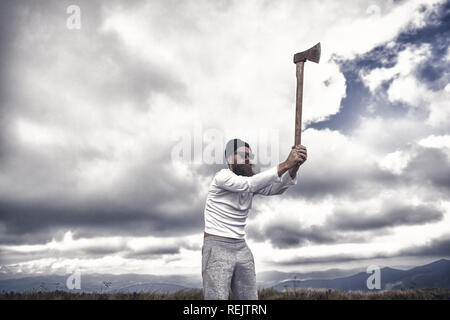 lumberjack. Hipster bearded lumberjack in sunglasses, hat raise axe on mountain landscape on cloudy sky. Logging and chopping concept. Wanderlust, hiking, vacation, travelling. Stock Photo