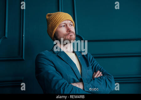 Attractive adult bearded angry man hipster in yellow hat looks at camera and furrowed his brows on blue wall background Stock Photo