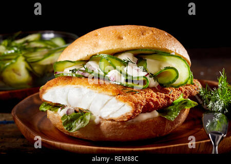 Fish burger with crumbed fillet of fish and sliced cucumber on a crusty bun served on a plate on rustic wood background Stock Photo