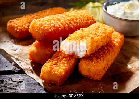 Crumbed golden fried fish fingers in sticks served on brown paper on a rustic wood counter with tartare sauce in a close up view with one broken open Stock Photo