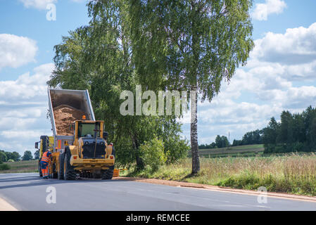 Road construction workers repairing highway road on sunny summer day. Loaders and trucks, heavy vibration roller compactor with arrow road sign on new Stock Photo