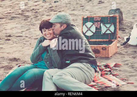 Couple in love first date having picnic outdoor smmiling and kissing eachother - middle age adults cacuasian people at the beach in winter enjoying wi Stock Photo