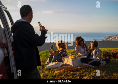 picnic and travel concept for family caucasian people enjoying the sunset sitting in the meadow and drinking beer together - children eating having fu Stock Photo