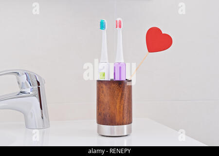 Two electric toothbrushes in wooden glass on sink in bathroom with red heart. Toothbrush for personal healthcare dental care. Oral dental hygiene. Den Stock Photo