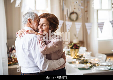 Senior couple standing indoors in a room set for a party, hugging.