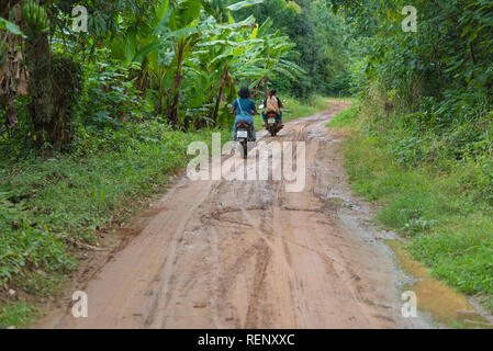 Nam Tok Sai Yok Noi, Thailand - August 18, 2018: Two scooters with three persons on them leave away along the dirt road into the green vegetation. Stock Photo