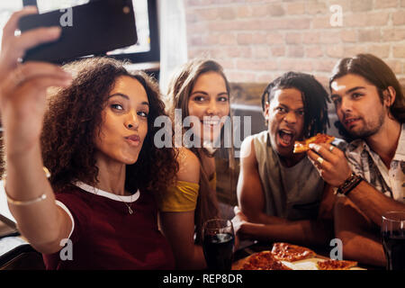 Woman taking selfie with friends eating pizza at a restaurant. Diverse group of men and women enjoying meal and taking selfie at restaurant. Stock Photo