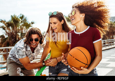 Group of three young friends having fun outdoors. Women with basketball and skateboard laughing with male friend. Stock Photo