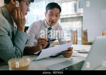 Young asian businessman explaining a document to his manager sitting at cafe table. Business professionals discussing over some paperwork at coffee sh Stock Photo
