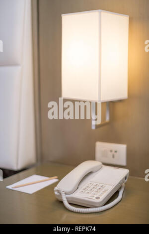 Hotel bedroom white Telephone on a bedside table with note pad and pencil a bedside lamp and plug socket all in a bright scene Stock Photo