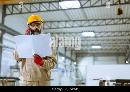 Handsome mature engineer looking at plan and using mobile phone in the factory Stock Photo