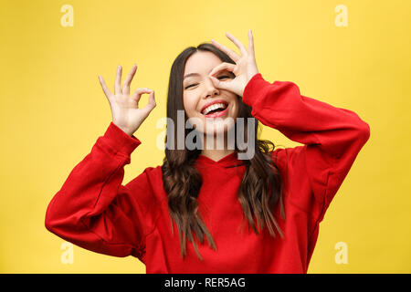 Close up portrait of beautiful joyful Caucasian female smiling, demonstrating white teeth, looking at the camera through fingers in okay gesture. Face expressions, emotions, and body language Stock Photo