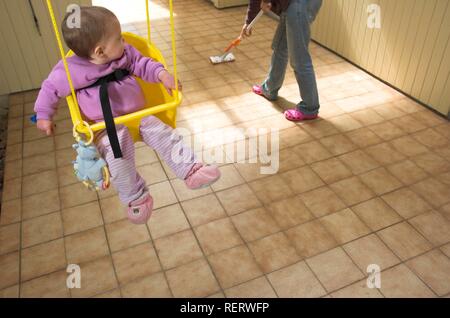 Baby girl in a swing looking over her shoulder at her mother mopping a tiled floor Stock Photo