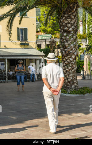 GARDONE RIVIERA, ITALY - SEPTEMBER 2018: Person with sun hat strolling around the town square in Gardone Riviera on Lake Garda with his hands clasped  Stock Photo
