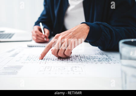 Close up of young architect designer in office working on notebook and building blueprint. Man engineer looking at drawing and computer while sits by  Stock Photo
