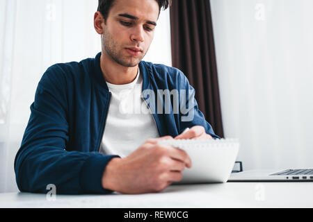 Bearded man using laptop and preparing project plan sitting at table his modern home. Concentrated man writing new ideas in notepad looking on screen  Stock Photo