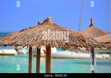 Moored boats float in crystal clear turquoise waters behind rustic parasol sunshades. A vision of a dream beach vacation. Stock Photo