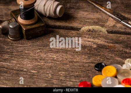 Close-up view of sewing spools with white and black thread and needle, thimbles and variety of colorful buttons set on wooden plank table Stock Photo