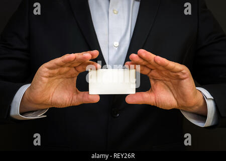 Man wearing a suit holding white business card on black wall background. Mock up, space for text Stock Photo