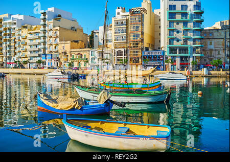 ST JULIANS, MALTA - JUNE 20, 2018: Spinola Bay harbor with colored wooden luzzu and modern motor boats, residential edifices and cafes along the prome Stock Photo