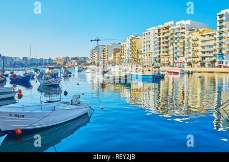ST JULIANS, MALTA - JUNE 20, 2018: The calm waters of Spinola Bay harbor reflect the moored colored boats and modern residential buildings along its s Stock Photo