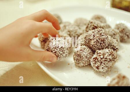 Close up of a white child hand picking  up a handmade chocolate and cocos sweet cookie bullet from a plate Stock Photo