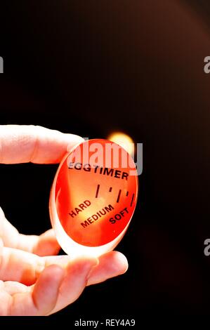 Red plastic egg timer held in white hand, on black background and back lighted. Used for hard, soft or medium boiled egg Stock Photo