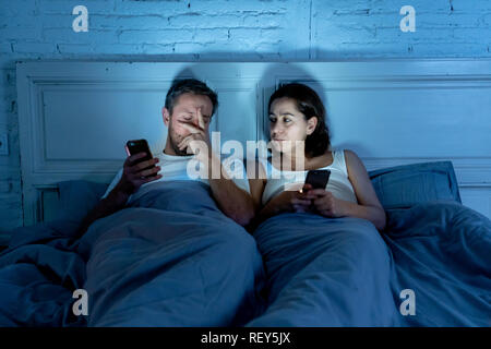 Couple affected by technology addiction ignoring at each other in apathy and anger lying in bed using their smart phones late at night in darkness in  Stock Photo