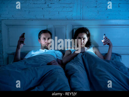 Couple affected by technology addiction ignoring at each other in apathy and anger lying in bed using their smart phones late at night in darkness in  Stock Photo