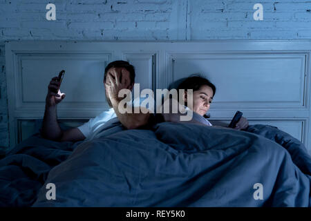 Couple affected by technology addiction ignoring at each other in apathy and anger lying in bed using their smart phones late at night in darkness in  Stock Photo