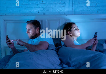 Couple affected by technology addiction ignoring at each other in apathy and anger lying in bed using their smart phones late at night in darkness in  Stock Photo