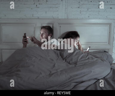 Couple affected by technology addiction ignoring at each other in apathy and anger lying in bed using their smart phones late at night in darkness in  Stock Photo