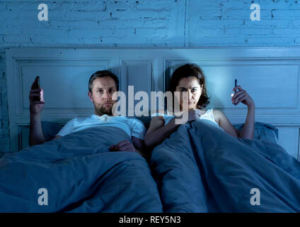 Couple affected by technology addiction ignoring at each other in apathy and anger lying in bed using their smart phones late at night in darkness in  Stock Photo