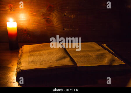 Reading scene in ancient times: an old book leaning on ruined wooden table lighted by a candle on a wooden background Stock Photo