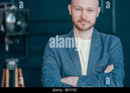 Portrait of attractive adult successful bald bearded man in suit on blue background, blogging, TV host