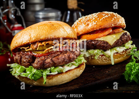 Two delicious beef burgers with salad trimmings, one a cheeseburger and the other with ham and ketchup on a dark wooden board in a close up view Stock Photo