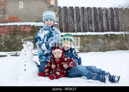 Little cute smiling baby boy and his two older brothers, sitting outdoors in the snow, snowman next to them Stock Photo