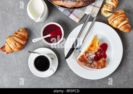 a portion of dutch baby topped with raspberry jam on a plate with fork and knife, croissants, cup of coffee and fresh cream on a concrete table, view  Stock Photo